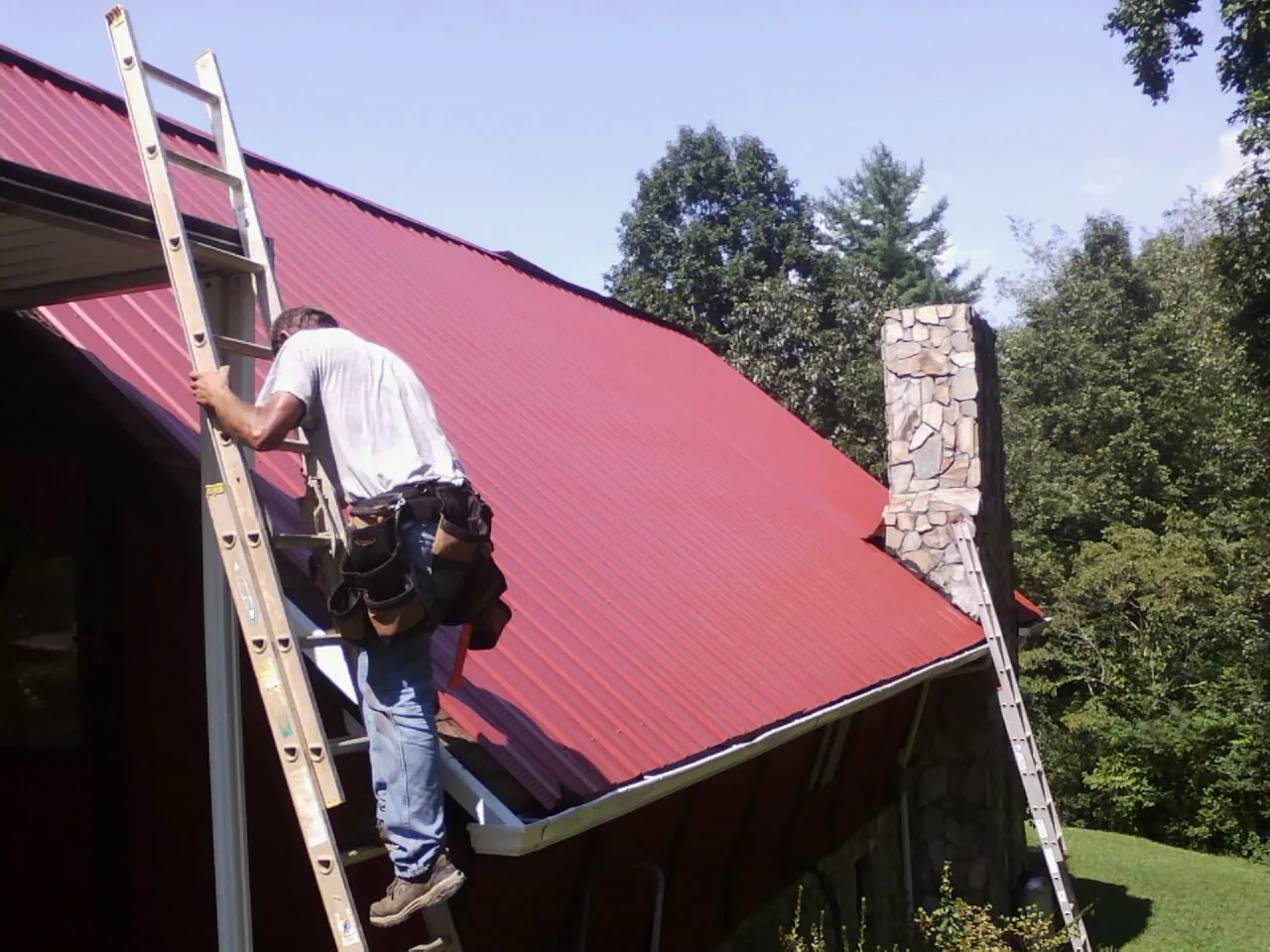 Roofer on a ladder beside a red metal roof and stone chimney — Soffit & Fascia Repair by Roofing Brothers in Osceola